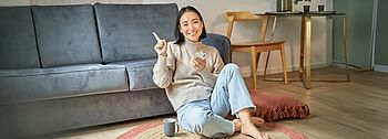 Nahwärme: Zufriedene und junge asiatische Frau sitz auf dem Boden im Wohnzimmer mit Handy in der Hand und einer Tasse neben sich.