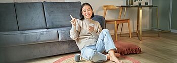 Nahwärme: Eine Option für nachhaltiges Heizen? Nahwärme: Zufriedene und junge asiatische Frau sitz auf dem Boden im Wohnzimmer mit Handy in der Hand und einer Tasse neben sich.