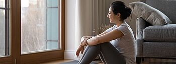 Kosten einer Lüftungsanlage: Junge Frau sitzt auf dem Fußboden in ihrem Wohnzimmer und blickt aus dem Fenster.