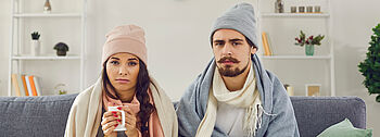 Heizung geht nicht – Was ist zu tun? Heizung geht nicht: Junges Paar sitzt frieren mit Tassen, Mützen und Decken auf einer Couch im Wohnzimmer.