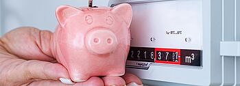 Heizkostenverordnung: Das müssen Sie wissen Heizkostenverordnung: Hand wirft münzen in ein rosanes Sparschwein, im Hintergrund ist ein Heizungszähler.