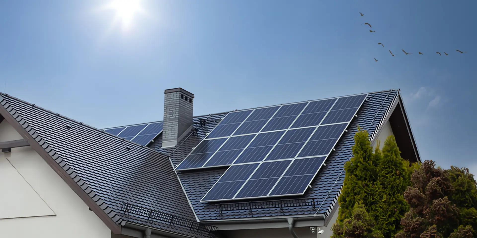 Einspeisevergütung Photovoltaik: Ein Haus samt Dach mit einer Photovoltaik-Anlage, im Hintergrund sind Himmel, Sonne und Vögel zu sehen.