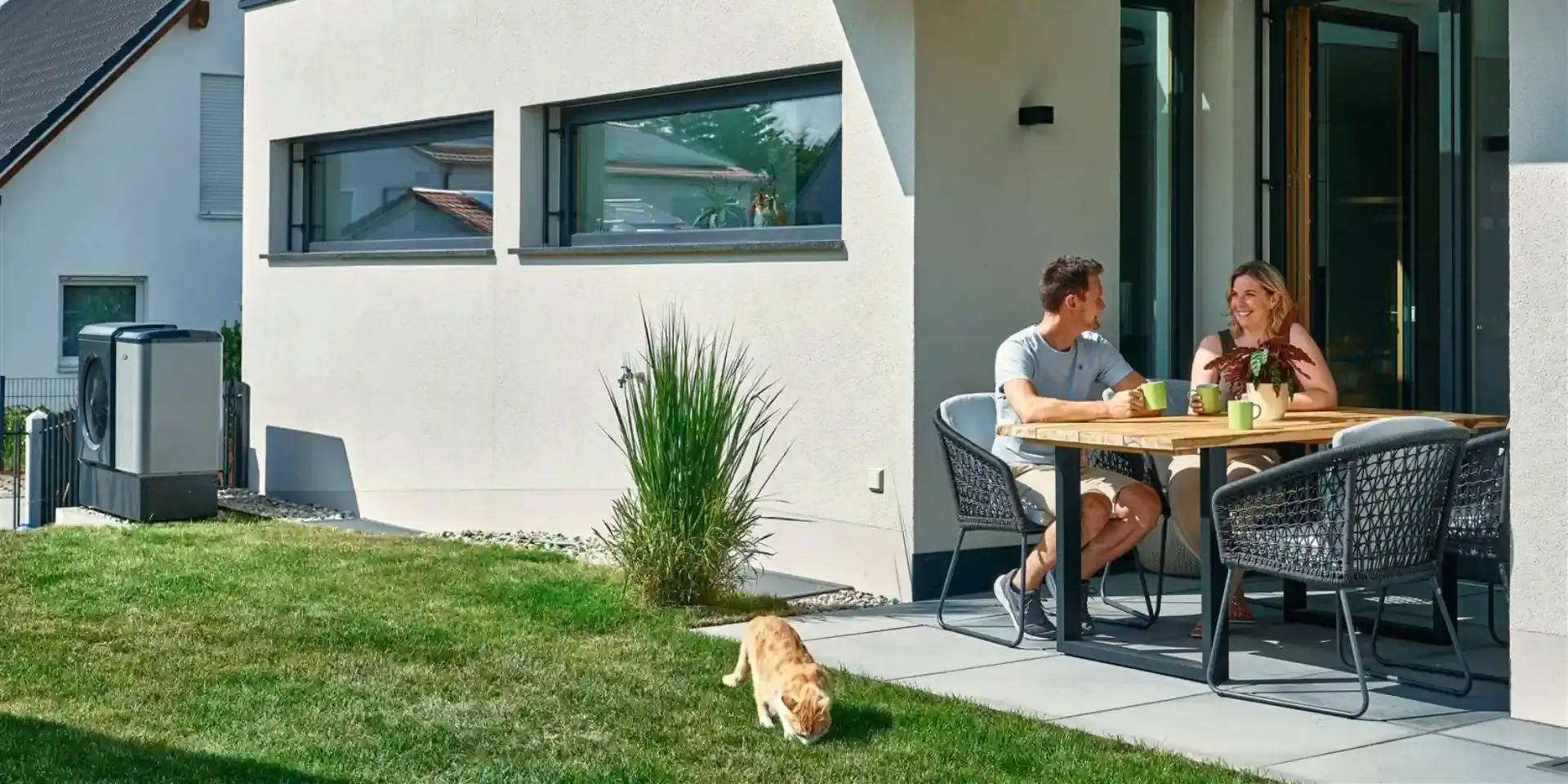 Heizung Beratung: Paar sitzt auf der Terrasse mit Wärmepumpe im Hintergrund